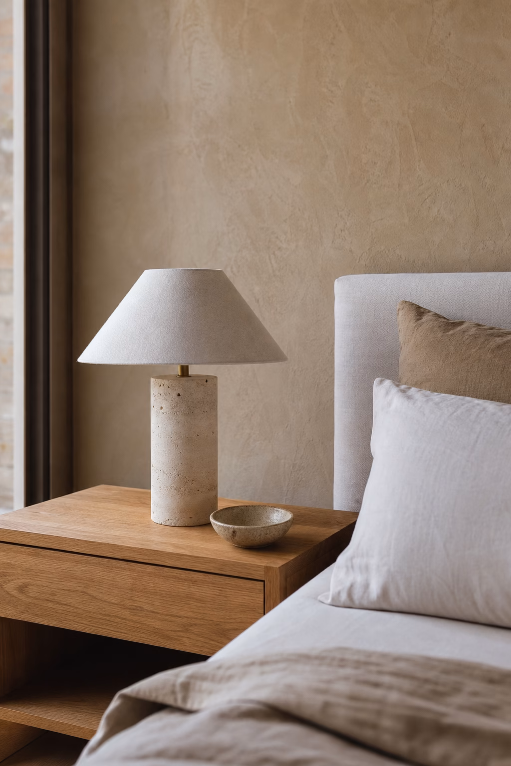 Earthy modern bedroom with travertine lamp, oak nightstand, linen bedding, and plaster wall