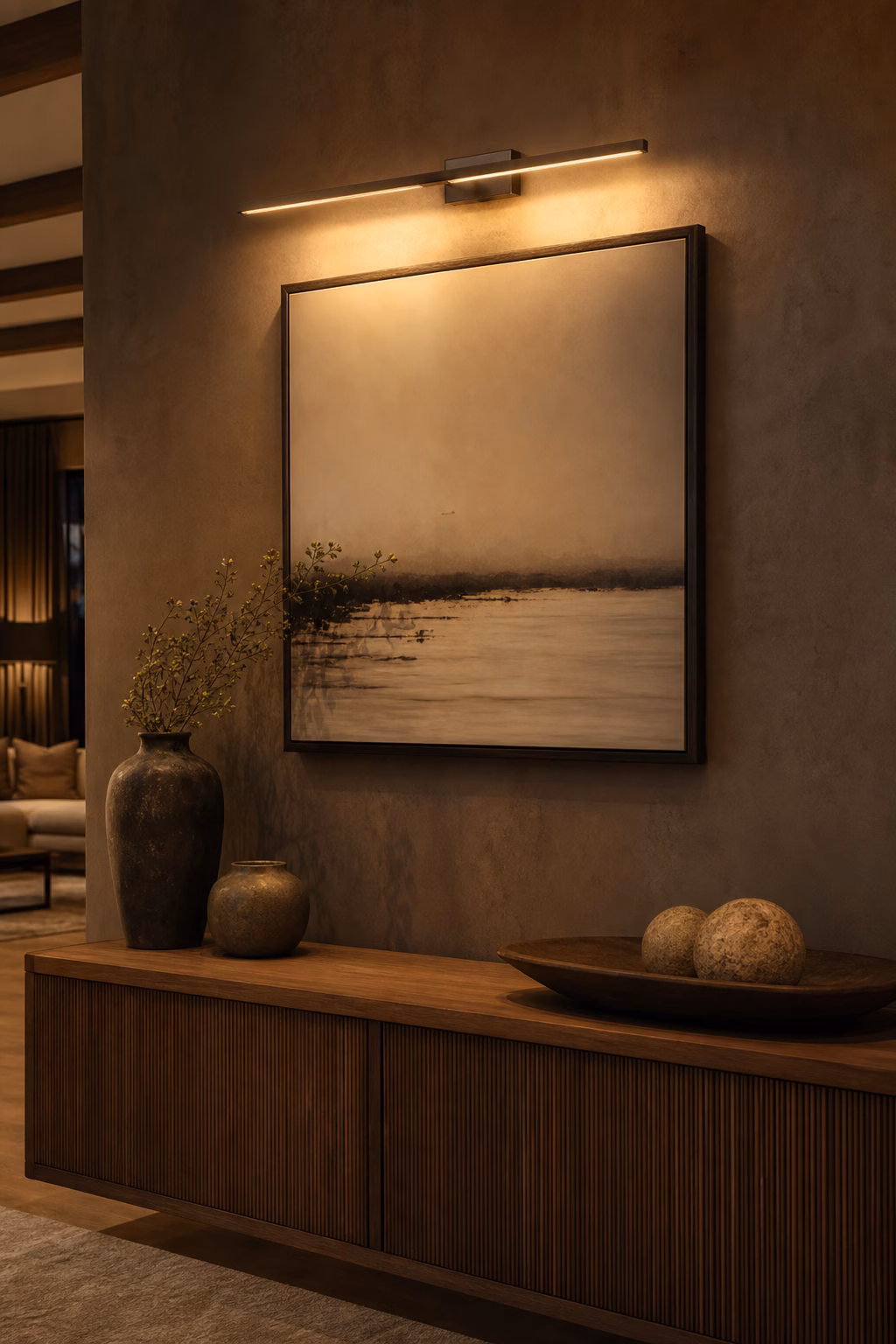 Artwork lit by a slim picture light above a console in a warm interior